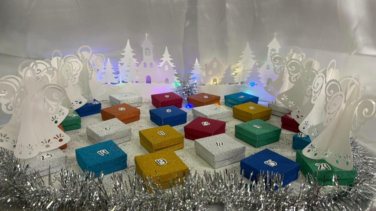 Calendrier de l'Avent de boîtes pailletées multicolores numérotées, entouré d'anges et d'un village enneigé en papier découpé, éclairés de guirlandes.