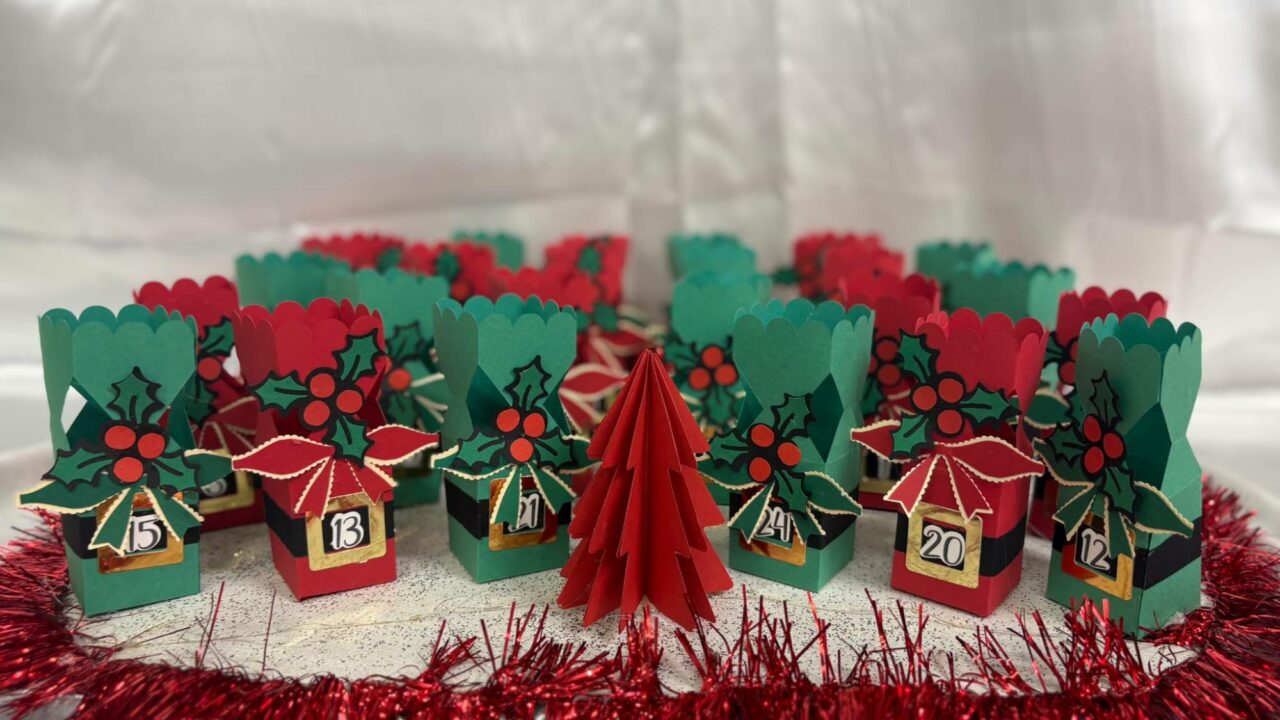 Calendrier de l'Avent de petites boîtes rouges et vertes en forme de bonhomme de Noël, décorées de houx, autour d'un sapin en papier rouge.