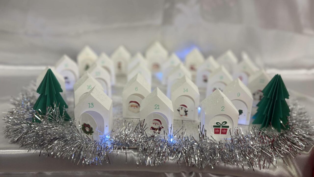 Calendrier de l'Avent de petites maisons blanches numérotées, avec une vignette de Noël sur chaque porte, entre deux sapins en papier vert.